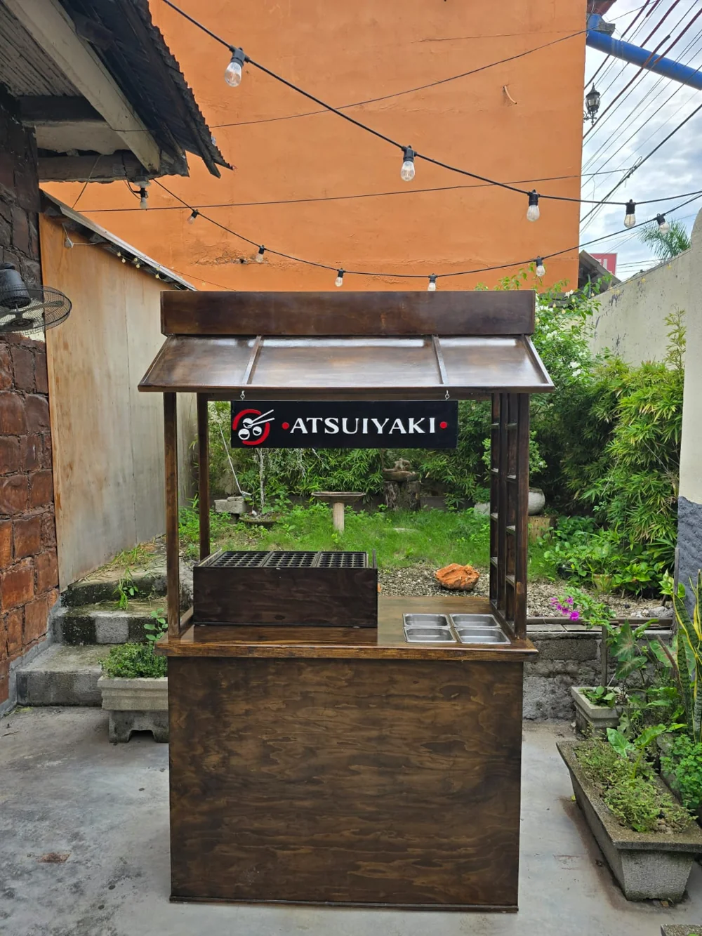 Puesto original de madera de Atsui Yaki, con su letrero, vegetación y guirnaldas de luces
